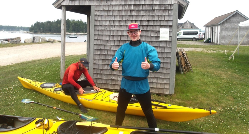 A person stands on green grass beside yellow kayaks and gives the camera two thumbs up